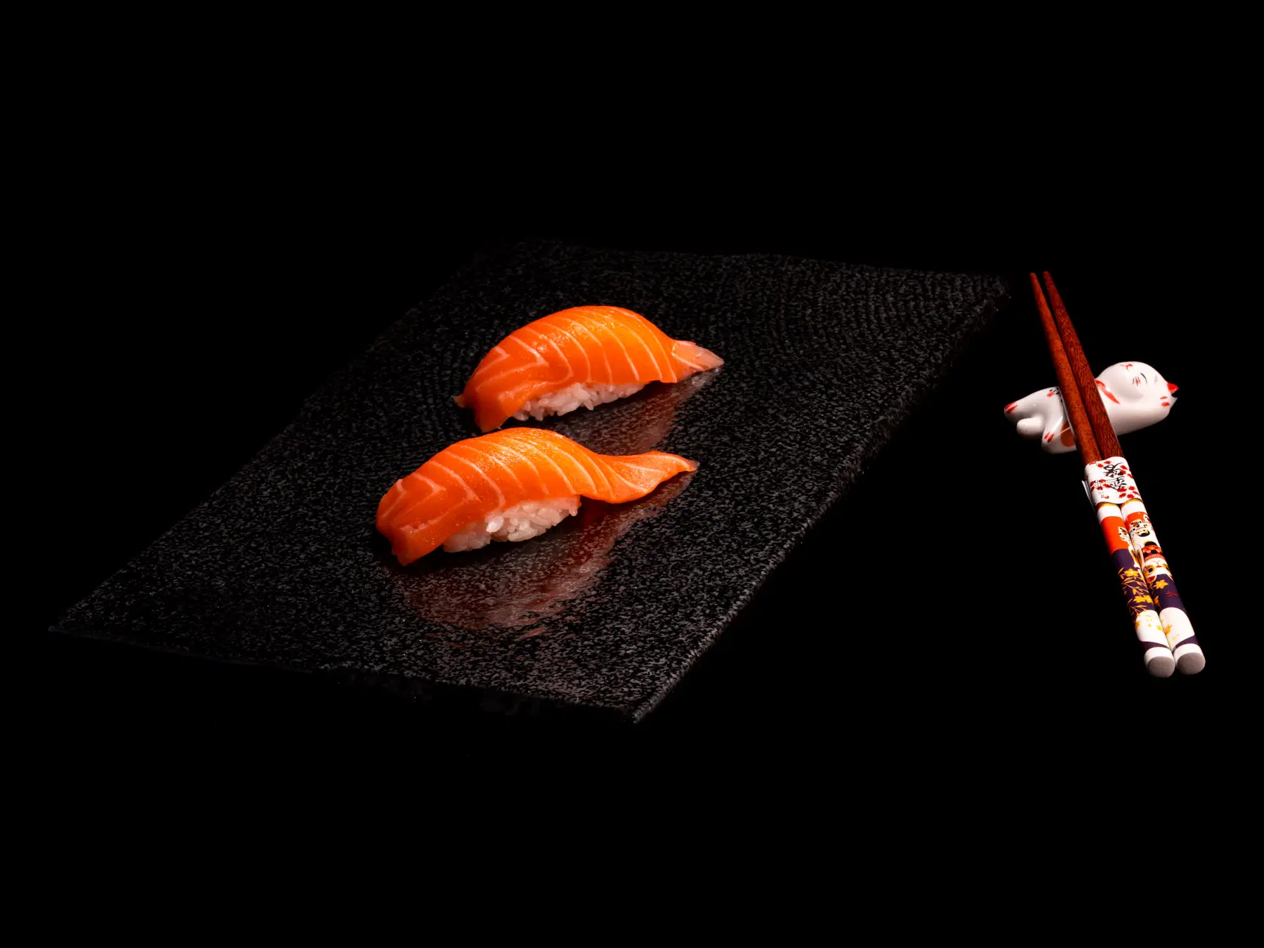 Two pieces of salmon nigiri on a dark stone slate beside a pair of chopsticks