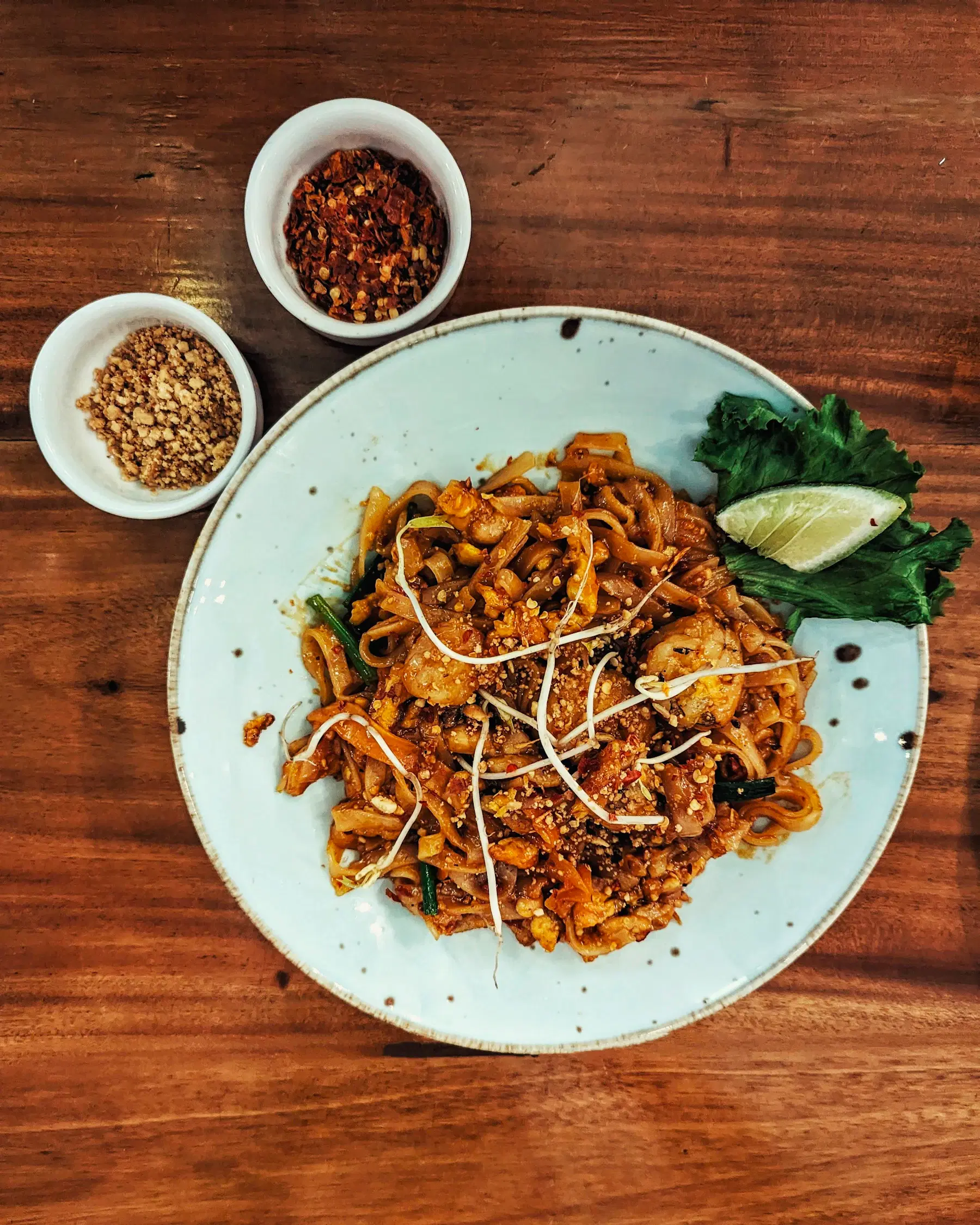 Pad Thai with shrimp, bean sprouts and lime on a speckled plate, with chili flakes and crushed peanuts on the side
