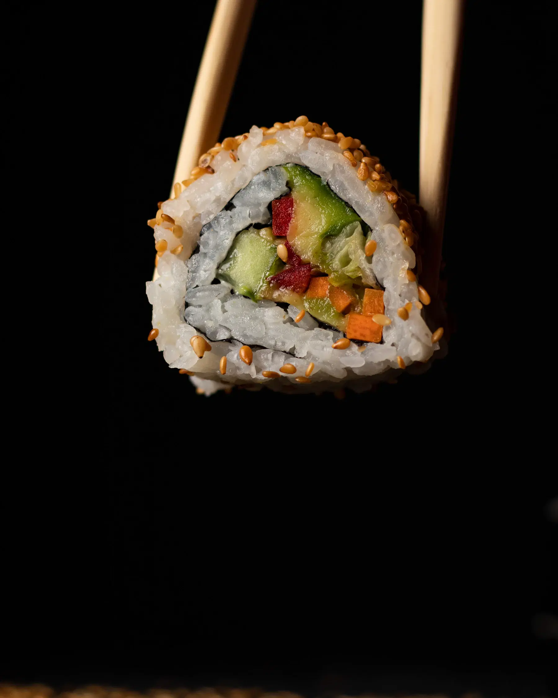 A vegetable sushi roll with avocado, cucumber, red pepper and carrot, coated in sesame and held in chopsticks
