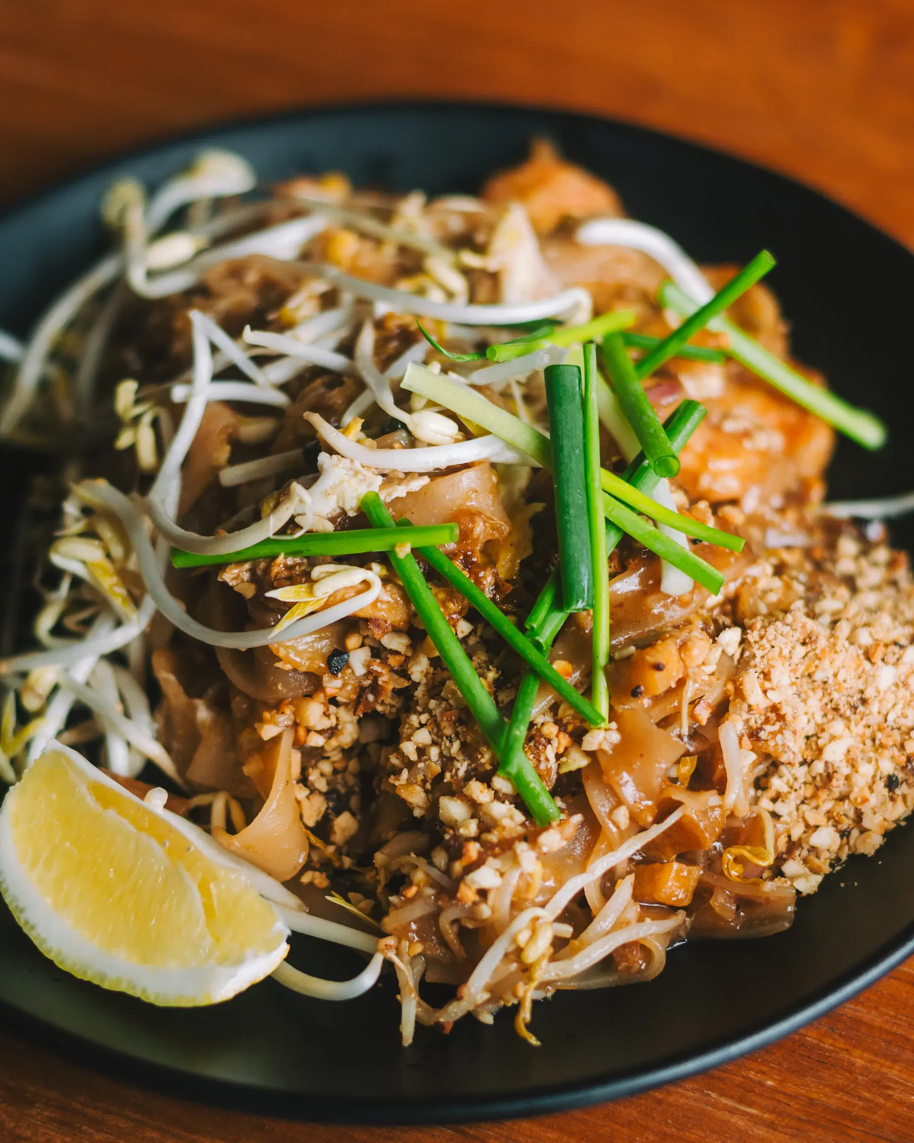 Pad Thai with rice noodles, bean sprouts, chives, crushed peanuts and a lemon wedge on a black plate